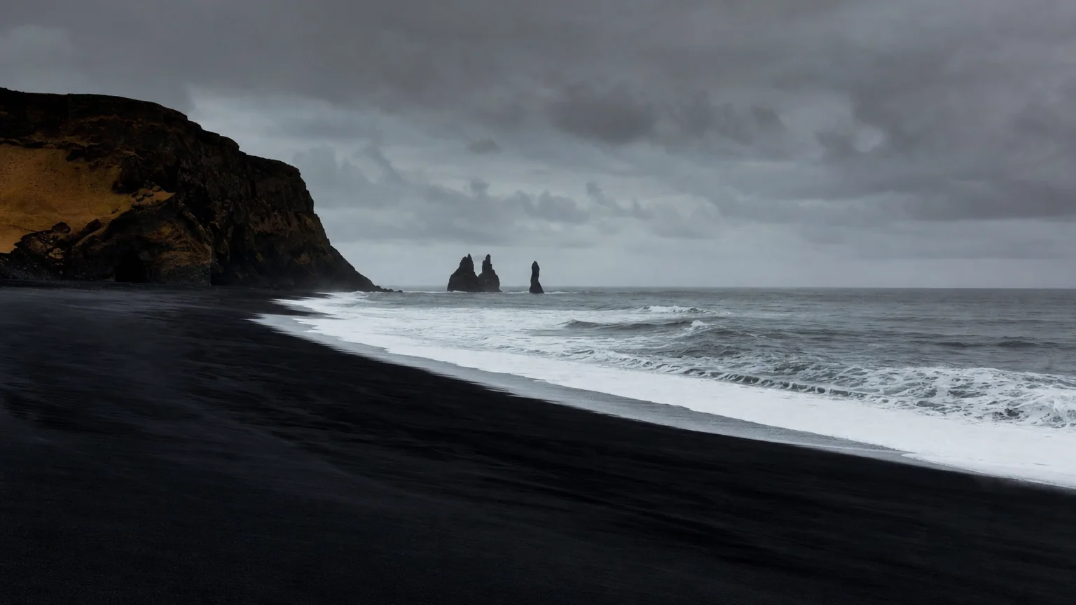 Black sand beach under a stormy sky, waves breaking against sea stacks